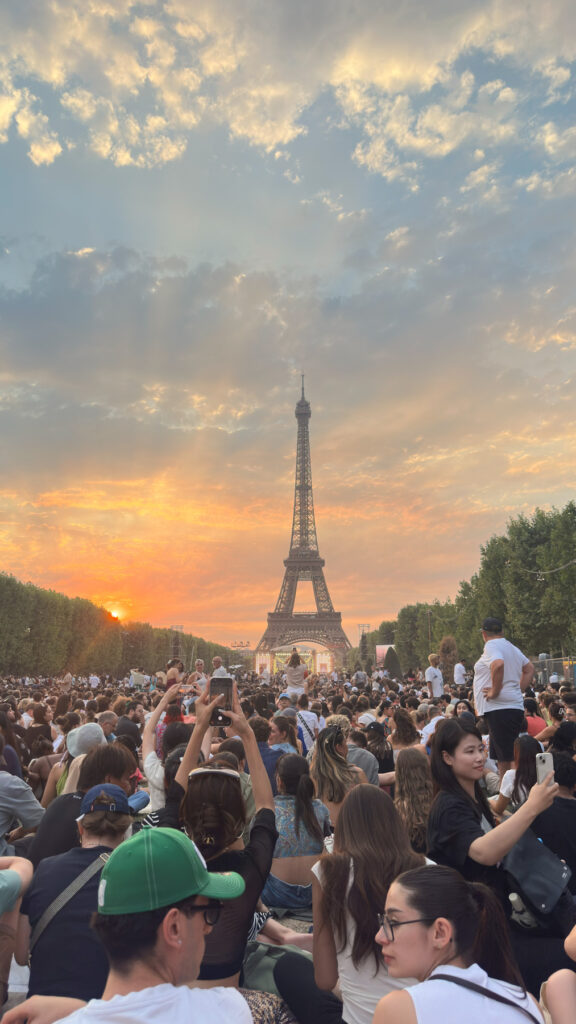 Une foule dense rassemblée au Champ-de-Mars face à la tour Eiffel au coucher du soleil, de nombreuses personnes photographiant la scène avec leur téléphone