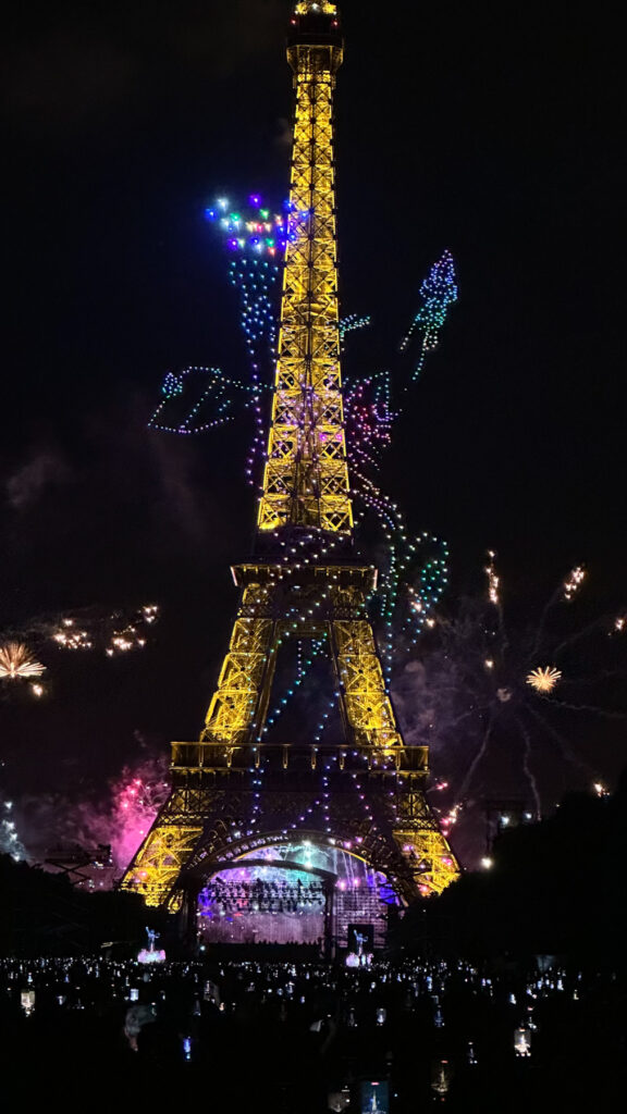 La tour Eiffel illuminée la nuit avec un spectacle de drones et de feux d'artifice colorés autour d'elle, devant une foule nombreuse