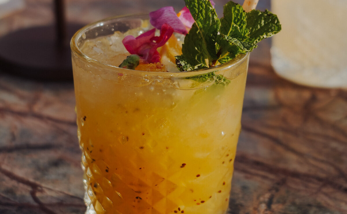Cocktail orangé sur glace pilée, garni de menthe fraîche et de pétales de fleur, servi sur une table en marbre