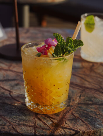 Cocktail orangé sur glace pilée, garni de menthe fraîche et de pétales de fleur, servi sur une table en marbre