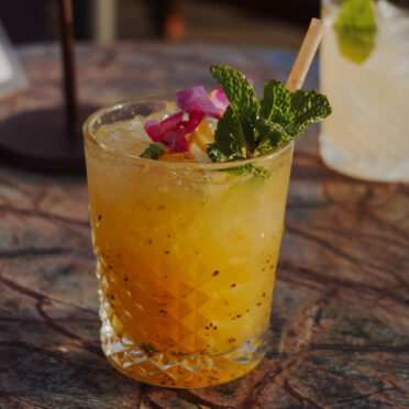 Cocktail orangé sur glace pilée, garni de menthe fraîche et de pétales de fleur, servi sur une table en marbre
