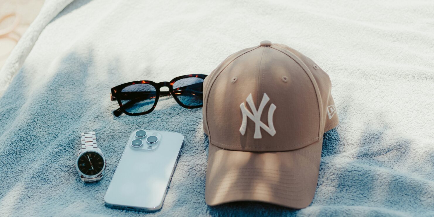 Casquette New York Yankees beige, lunettes de soleil, montre et smartphone posés sur une serviette de plage bleue