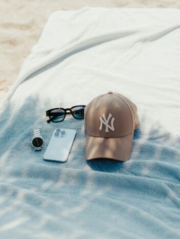 Casquette New York Yankees beige, lunettes de soleil, montre et smartphone posés sur une serviette de plage bleue