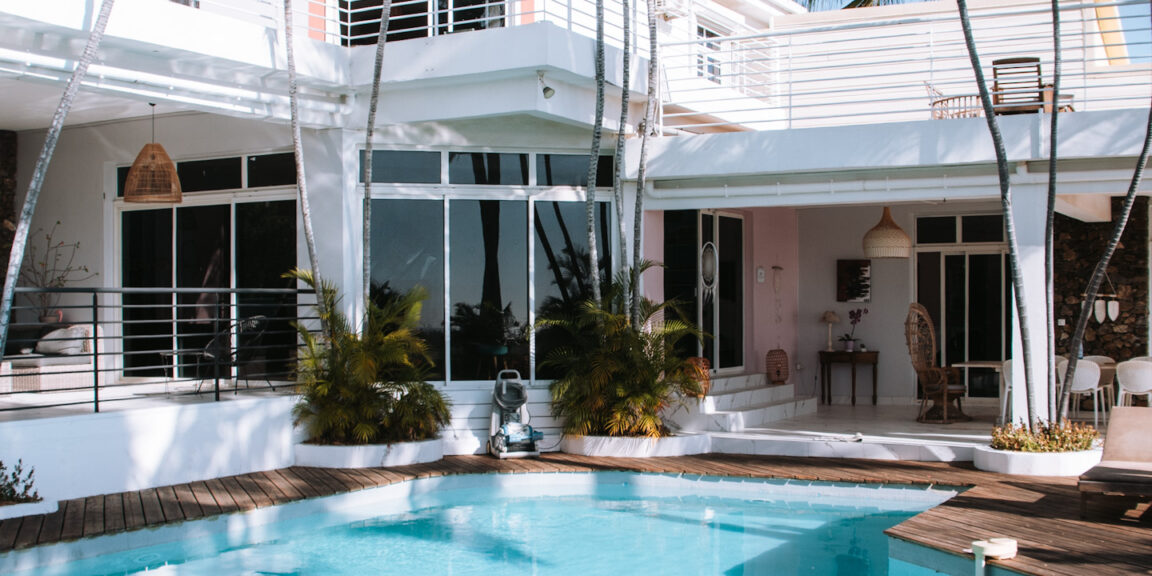 Villa moderne blanche avec piscine turquoise, terrasse en bois et palmiers sous un ciel bleu