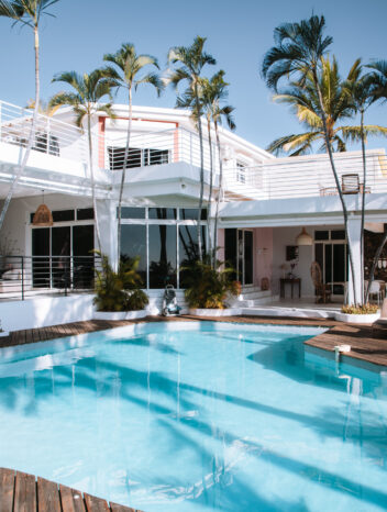 Villa moderne blanche avec piscine turquoise, terrasse en bois et palmiers sous un ciel bleu