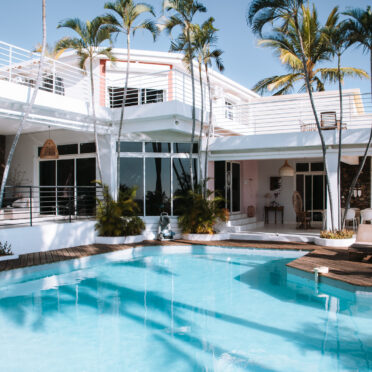 Villa moderne blanche avec piscine turquoise, terrasse en bois et palmiers sous un ciel bleu