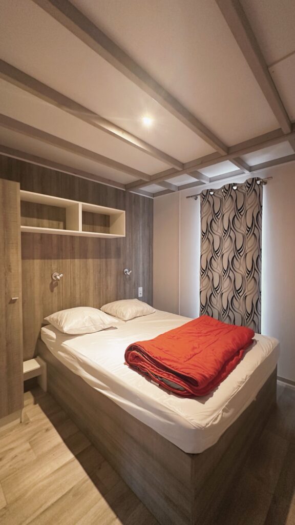 Chambre de mobil-home avec lit double, couverture pliée, rangements muraux et rideau à motifs