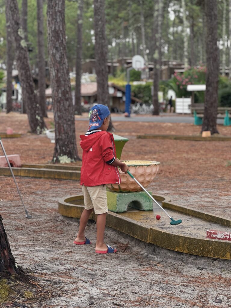 Enfant en veste rouge jouant au mini-golf sur un parcours ombragé par des pins au camping Eurosol