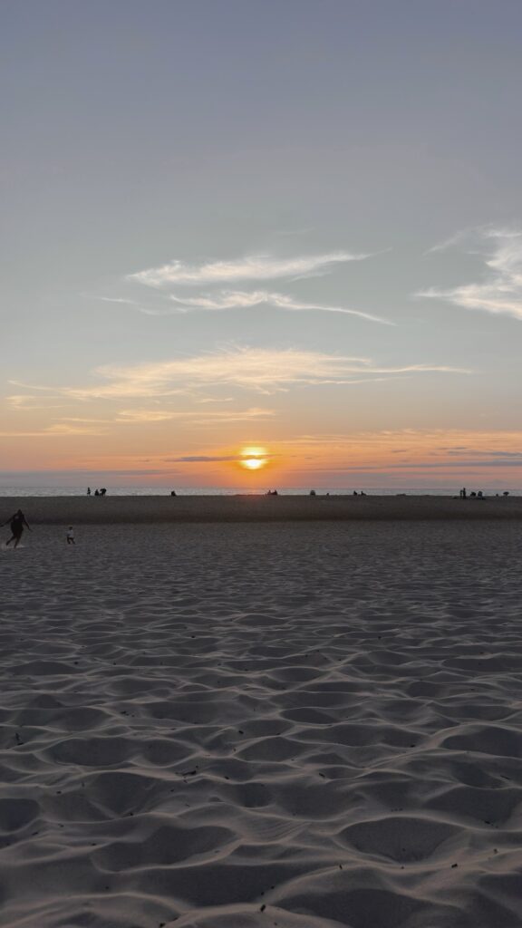 Coucher de soleil sur une plage, avec des silhouettes de promeneurs au bord de l'eau et le sable texturé au premier plan