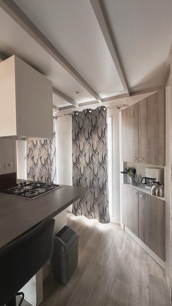 Cuisine équipée de mobil-home avec plaques de cuisson, meubles en bois clair et rideaux à motifs