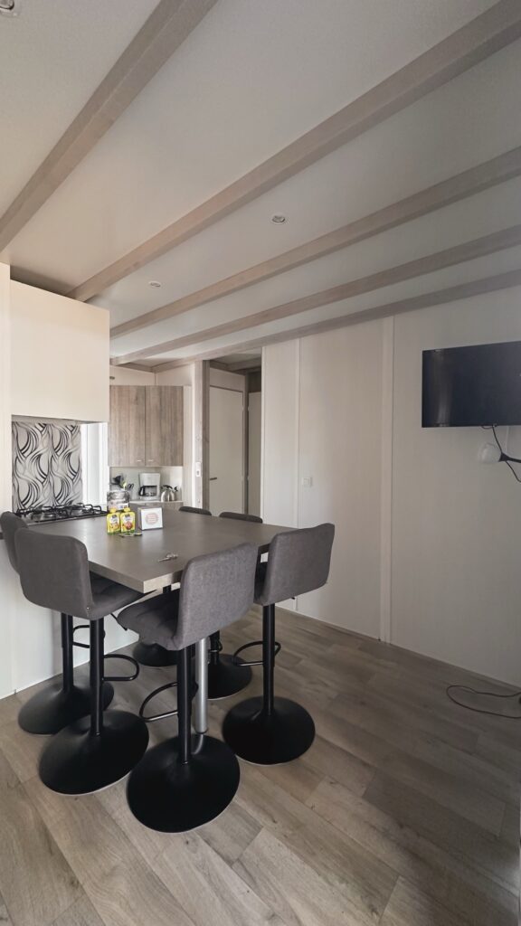 Cuisine ouverte avec table haute, six chaises grises, plaque de cuisson et télévision murale