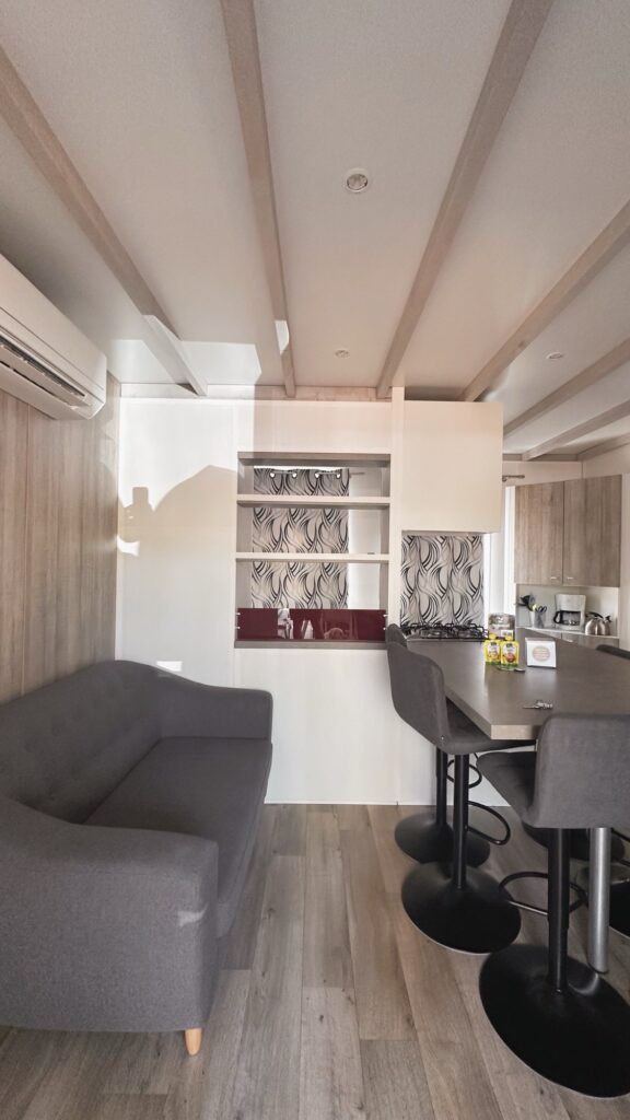 Salon et coin cuisine d'un mobil-home avec canapé gris, bar et tabourets, plaque de cuisson et rangements