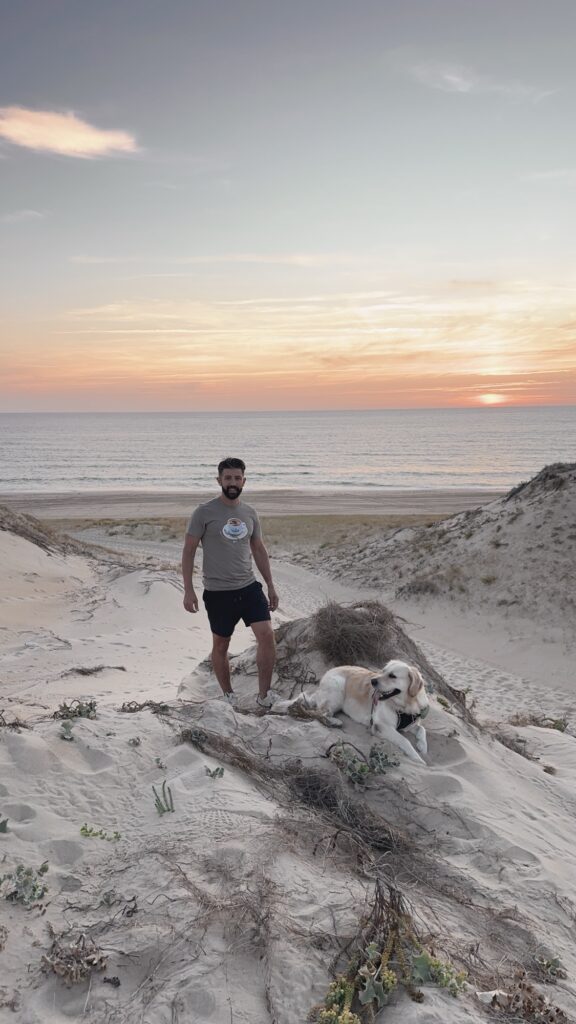 Homme debout sur une dune avec un golden retriever couché à côté, coucher de soleil sur la mer en arrière-plan