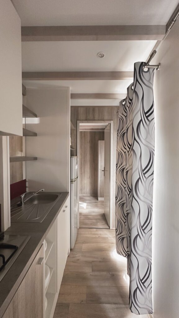 Cuisine étroite avec évier, réfrigérateur et couloir menant aux chambres, rideau à motifs abstraits en premier plan