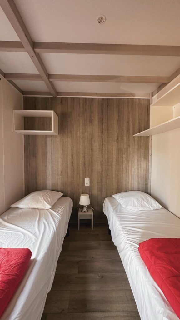Chambre avec deux lits jumeaux séparés par une table de chevet, linge blanc et rouge, rangements muraux
